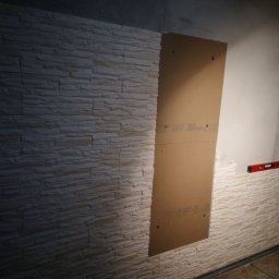 Grzegorz Kowalik - Montaż dekoracyjnej okładziny ściennej imitującej kamień, z częściowo zamontowaną płytą na ścianie i poziomicą dla wyrównania.