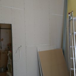 Grzegorz Kowalik - Ściana wykończona płytami, widoczne łączenia, metalowe profile, drabina, butelka wody i płyty leżące na podłodze w trakcie prac.