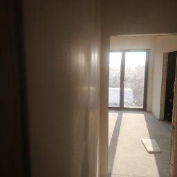 Grzegorz Kowalik - Świeżo nałożona, gładka ściana w jasnym odcieniu, z widocznymi refleksami światła słonecznego, w tle okno z widokiem na zewnątrz i fragment pomieszczenia w stanie surowym.