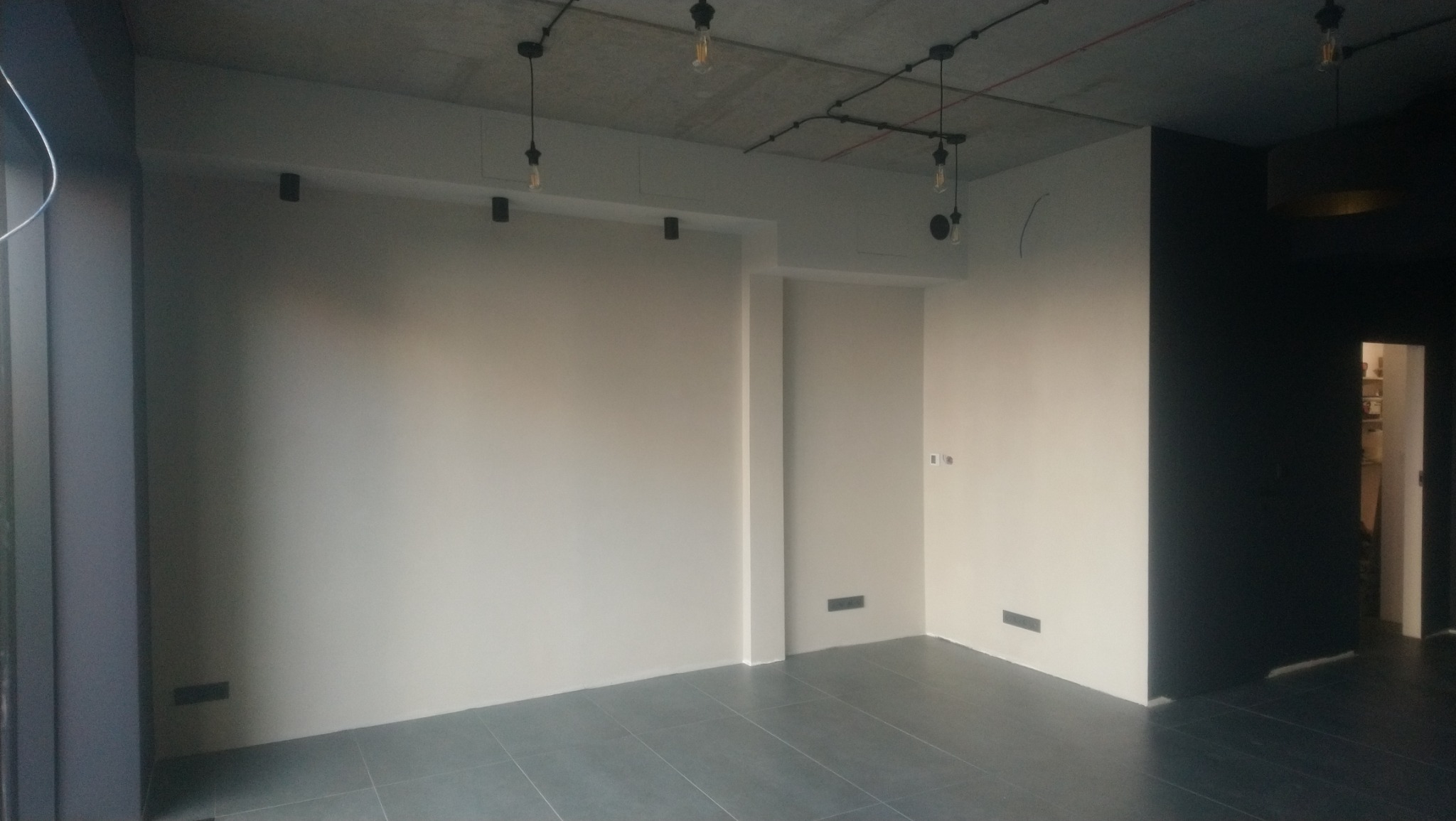 Wnętrze po szpachlowaniu ścian, z widocznym surowym betonowym sufitem z industrialnym oświetleniem i szarą podłogą z płytek.