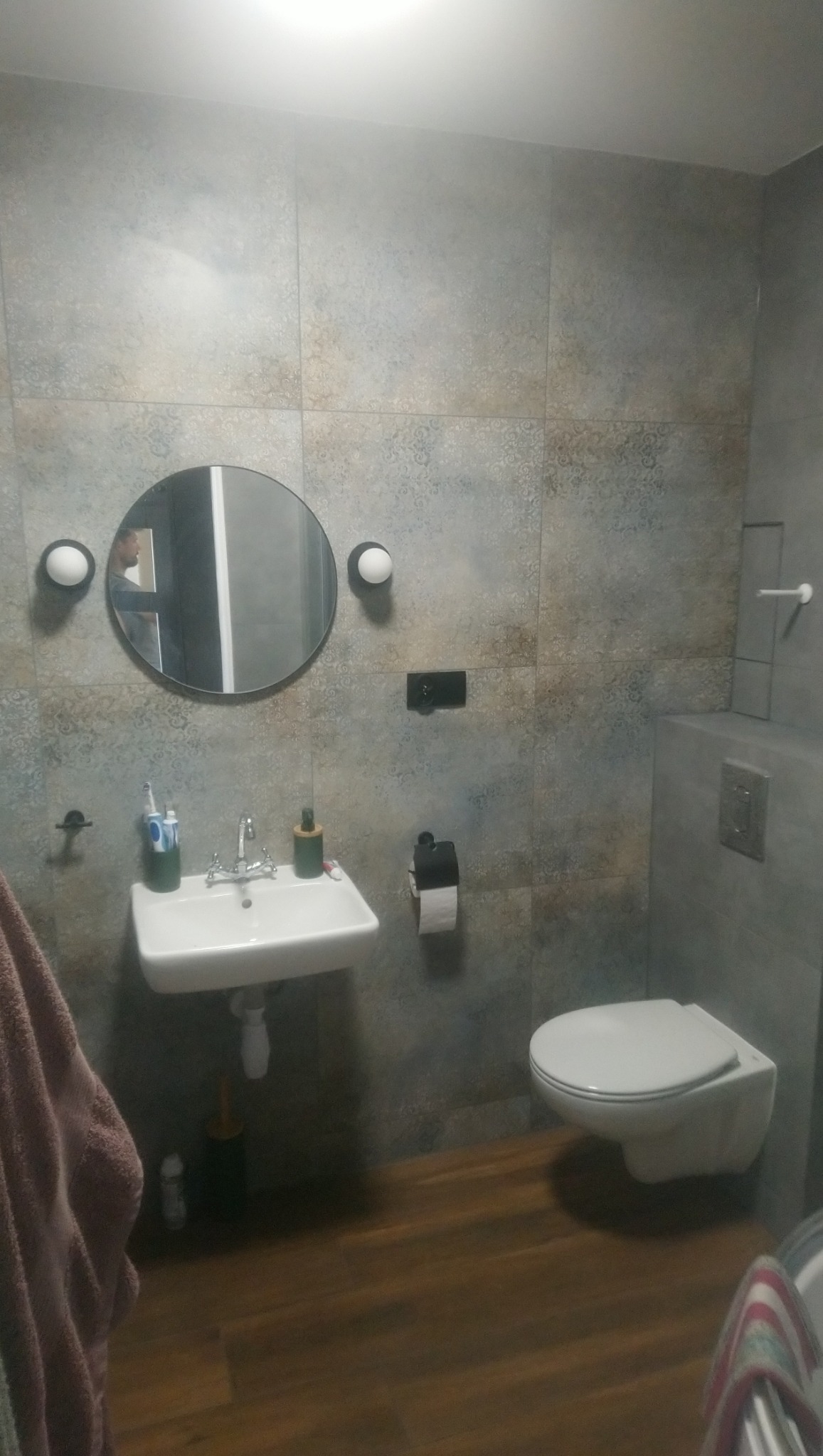 Nowoczesna łazienka z okrągłym lustrem podświetlonym kulistymi lampami, wiszącą umywalką i toaletą w zabudowie, wykończona płytkami w odcieniach szarości i brązu oraz podłogą imitującą drewno.