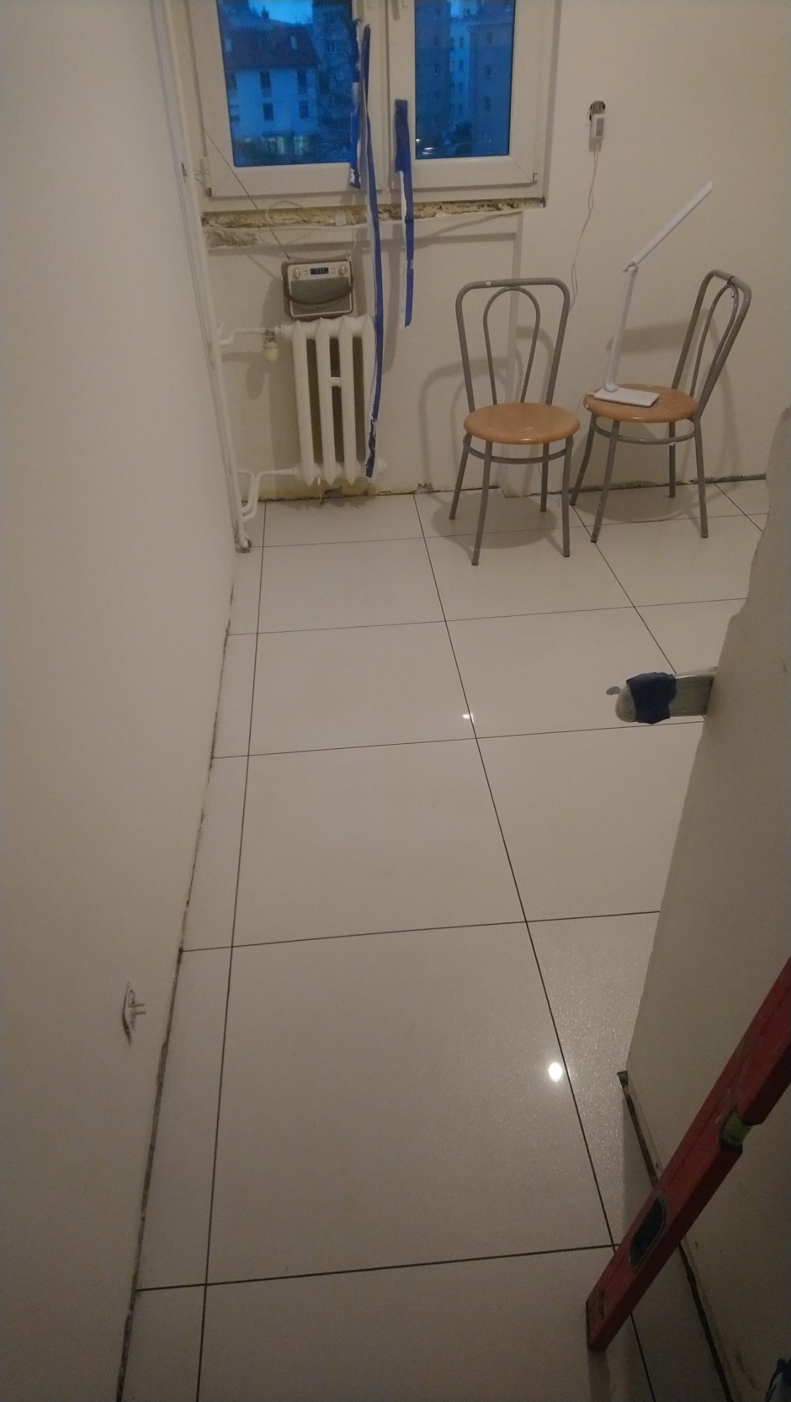 Świeżo ułożone, białe, błyszczące płytki podłogowe w trakcie remontu, widoczne narzędzia i poziomica przy ścianie, dwa krzesła i lampa w tle, zabezpieczenia okna folią malarską.