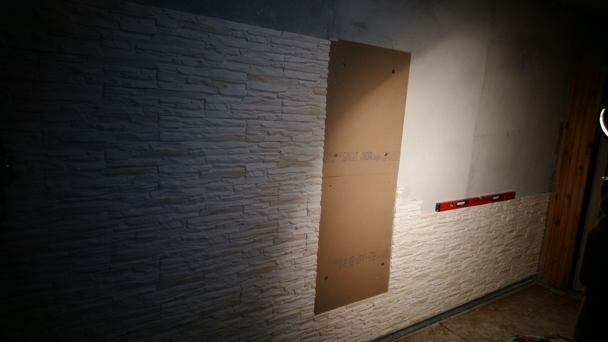 Montaż dekoracyjnej okładziny ściennej imitującej kamień, z częściowo zamontowaną płytą na ścianie i poziomicą dla wyrównania.