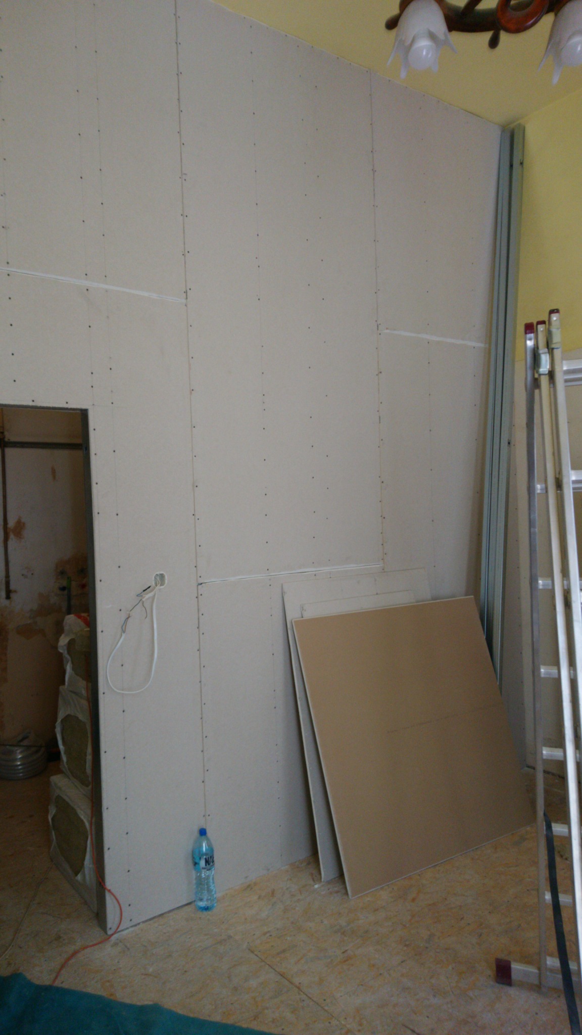 Ściana wykończona płytami, widoczne łączenia, metalowe profile, drabina, butelka wody i płyty leżące na podłodze w trakcie prac.