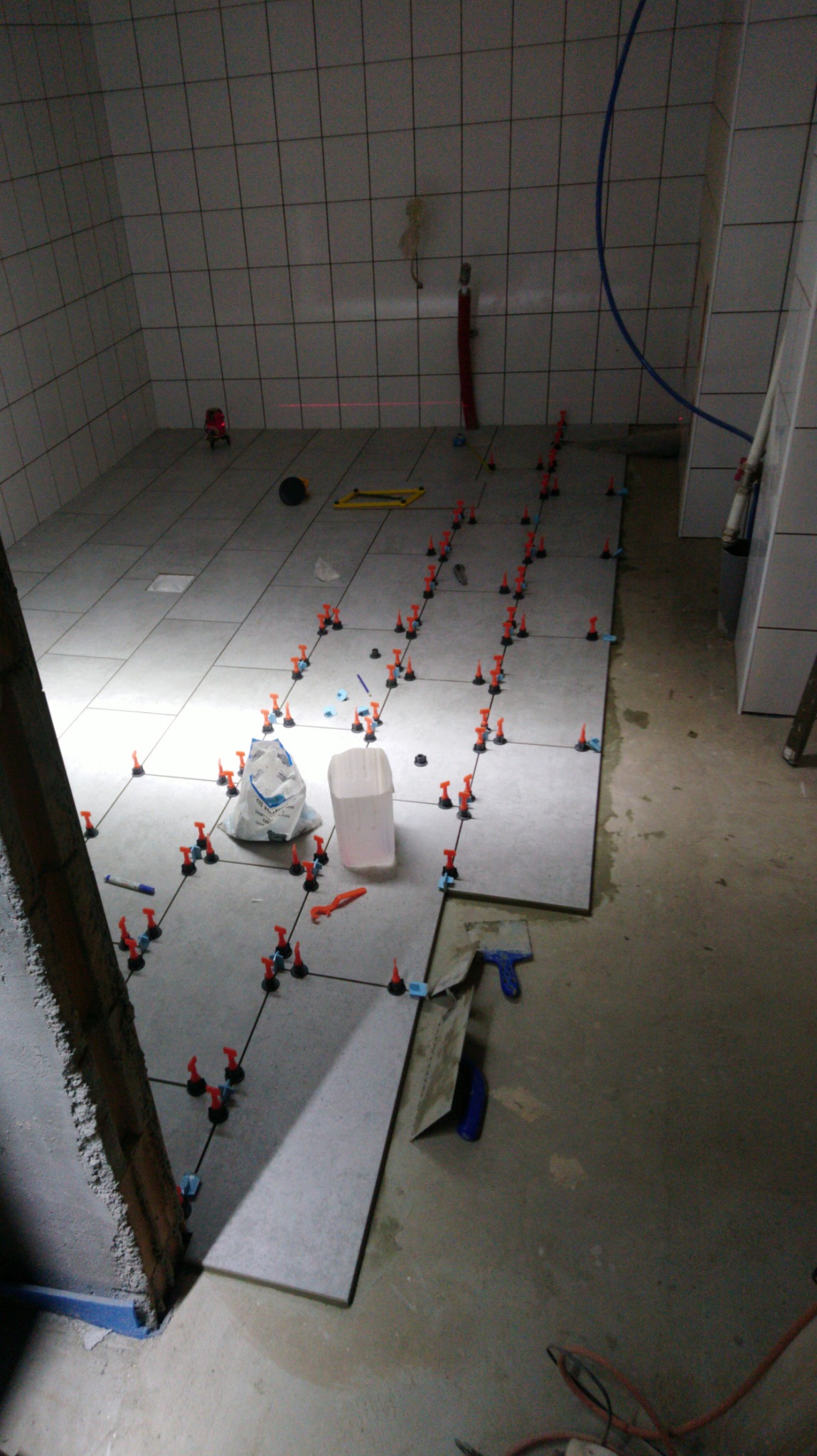 Układanie szarych płytek podłogowych w łazience, widoczne poziomowanie laserowe, system poziomowania płytek z pomarańczowymi klinami i niebieskimi bazami, białe wiadro i worek z zaprawą w tle.