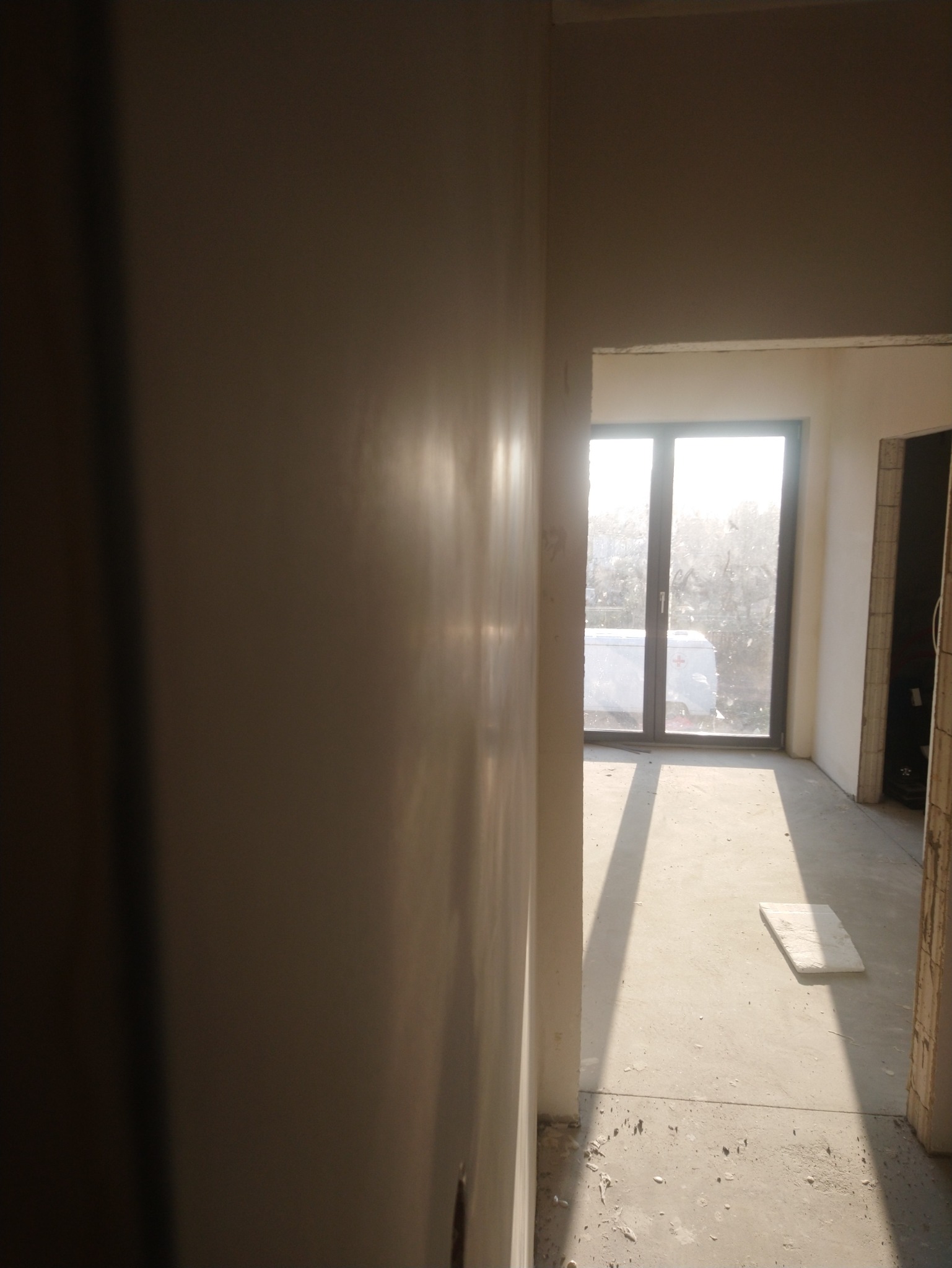 Świeżo nałożona, gładka ściana w jasnym odcieniu, z widocznymi refleksami światła słonecznego, w tle okno z widokiem na zewnątrz i fragment pomieszczenia w stanie surowym.