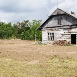 Niwelacja terenu przed drewnianym, częściowo murowanym domem z stertą drewna pod oknem, widoczny samochód dostawczy