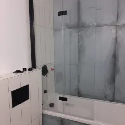 Nowoczesna łazienka z wanną i szklanym parawanem w odcieniach bieli i szarości, czarna armatura, płytki imitujące beton na ścianie w strefie kąpieli, widoczny fragment toalety.