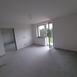 D&M Solutions - Wnętrze w stanie deweloperskim z surowymi ścianami i podłogą, widoczne wyprowadzenia instalacji elektrycznej na suficie i ścianach, otwarte okno balkonowe z widokiem na zieleń, lampa budowlana...