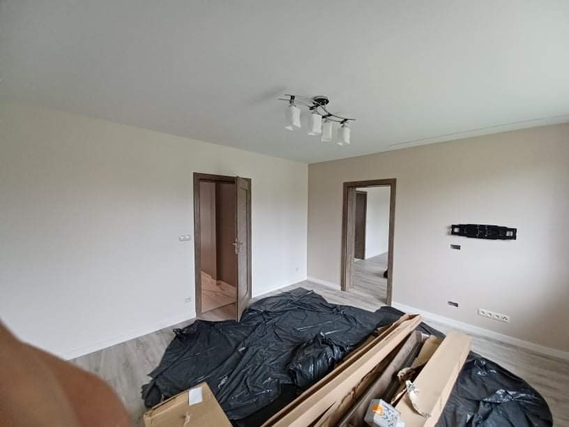 Pokój w trakcie remontu: świeżo pomalowane ściany w odcieniach beżu, zabezpieczona podłoga czarną folią, kartony z materiałami budowlanymi leżące na podłodze, widoczne ościeżnice drzwiowe w kolorze...