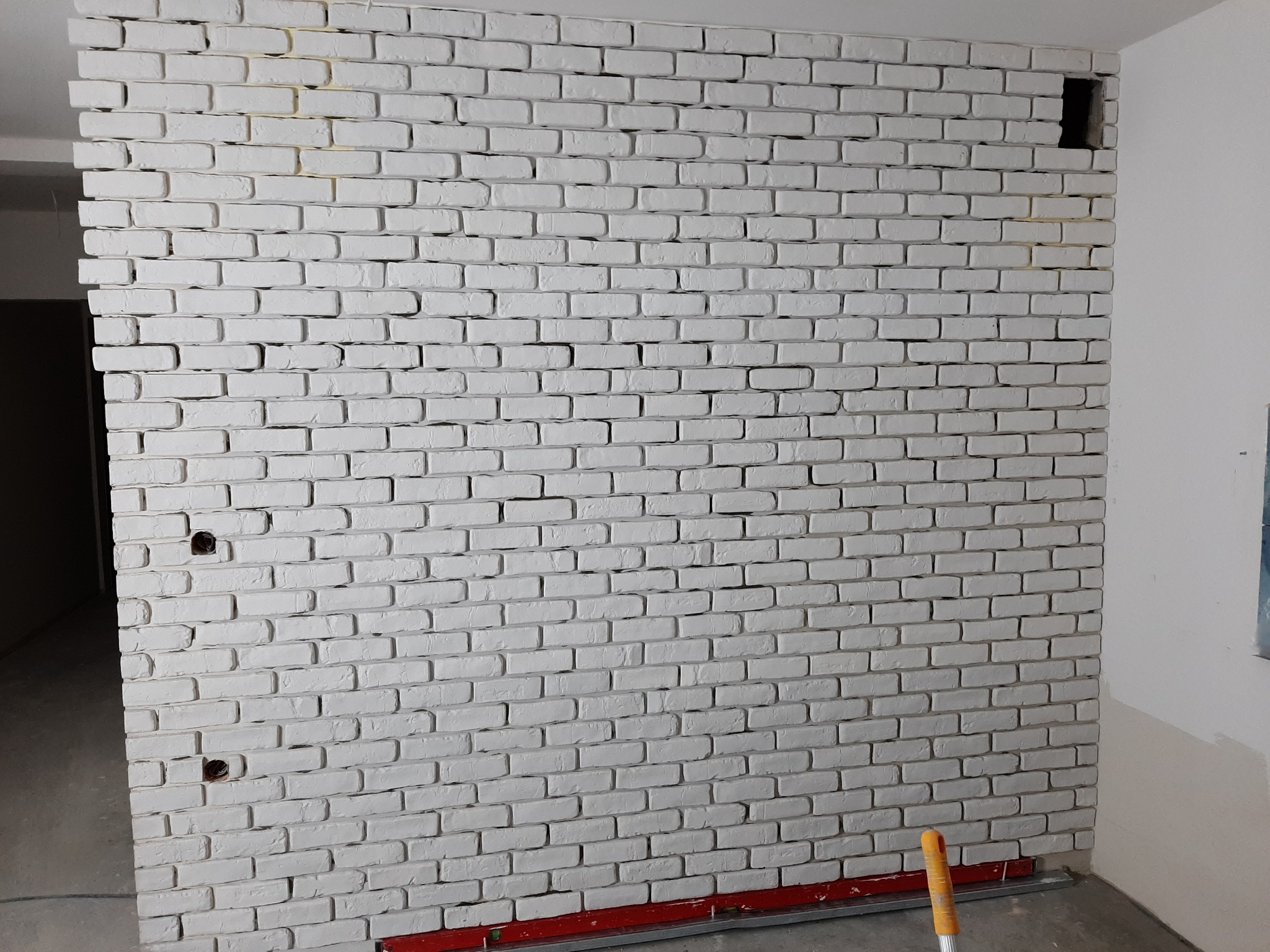 Ściana z białej cegły dekoracyjnej z widocznymi otworami na gniazdka elektryczne i miejscem na oświetlenie, na podłodze poziomica w kolorze czerwonym.