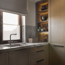 EMITOM design & build S.C. - Nowoczesna kuchnia z drewnianymi szafkami, marmurowym blatem i podświetlanymi półkami z dekoracyjnymi naczyniami, widok na okno z roletą rzymską.
