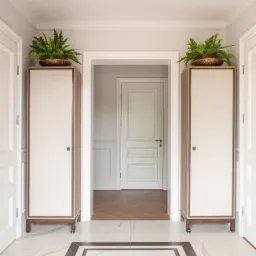 Elegancki przedpokój z marmurową podłogą w geometryczny wzór, szafkami z roślinami i widokiem na kolejne pomieszczenie z białymi drzwiami i panelami ściennymi.