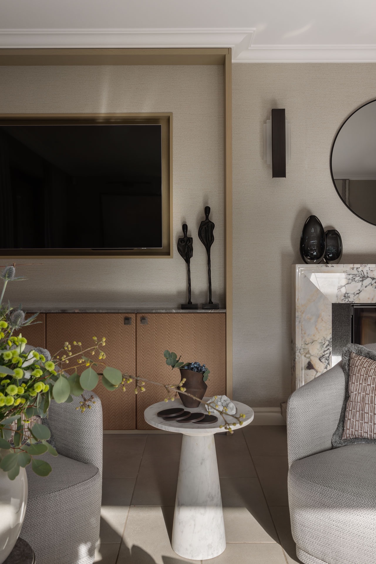 Elegancki salon z telewizorem w ramie, marmurowym kominkiem, szarymi fotelami i stolikiem z blatem z białego marmuru, dekoracjami i kwiatami, oświetlony nowoczesnymi kinkietami.