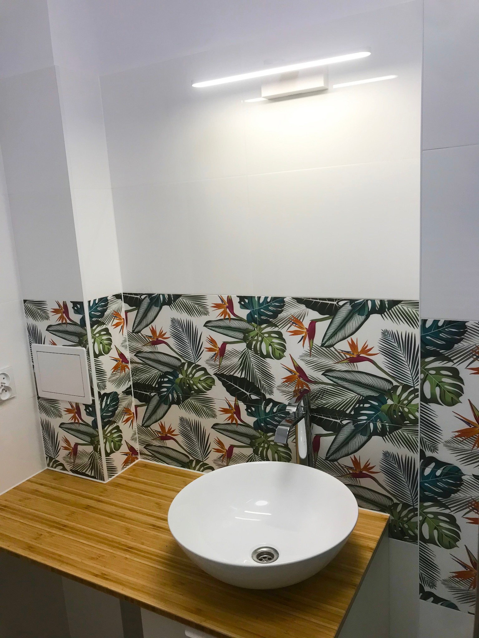 Biała, okrągła umywalka nablatowa na blacie z bambusowych listewek, tło stanowi ściana wyłożona płytkami z motywem tropikalnych liści i kwiatów strelicji, oświetlona podłużną lampą LED.