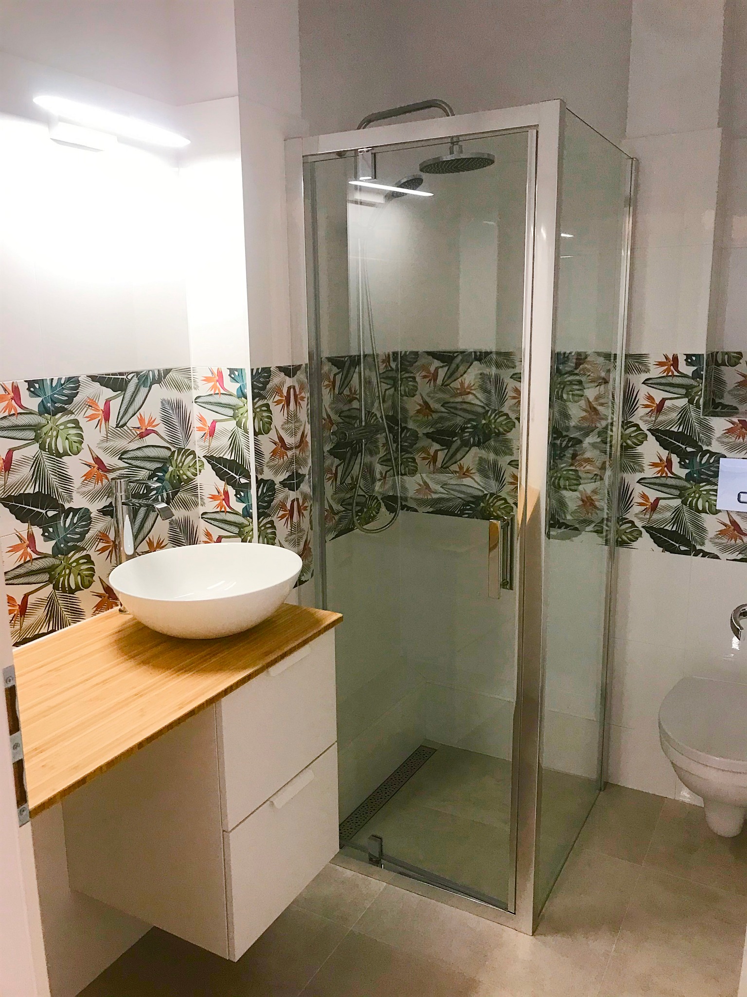 Nowoczesna łazienka z kabiną prysznicową, umywalką nablatową na drewnianym blacie i płytkami z motywem tropikalnym.