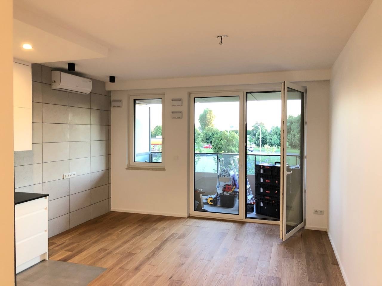 Jasne wnętrze z drewnianą podłogą, wykończoną ścianą z płytek imitujących beton, klimatyzatorem i otwartym balkonem z widocznymi narzędziami i skrzynkami.