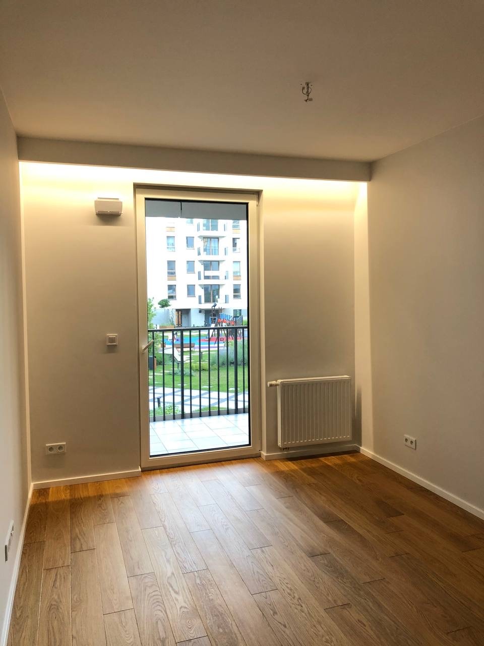 Wnętrze pokoju z drewnianą podłogą, balkonem z widokiem na zielony dziedziniec i oświetleniem LED pod sufitem.