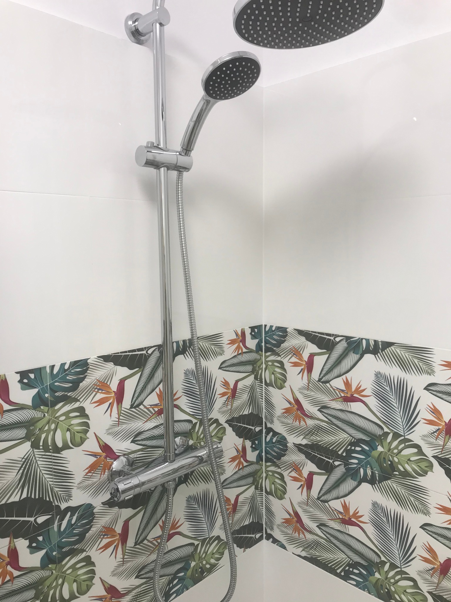 Bateria prysznicowa z dwoma słuchawkami, zamontowana na ścianie wyłożonej białymi płytkami oraz dekoracyjnymi płytkami z motywem tropikalnych liści i kwiatów strelicji.