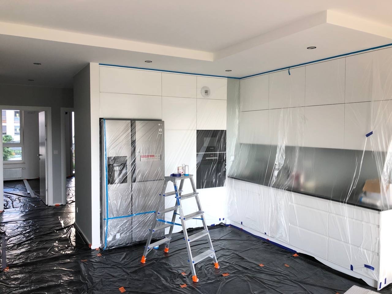 Przygotowanie kuchni do malowania: podłoga i meble zabezpieczone folią i taśmą malarską, drabina aluminiowa z puszką farby na tle zabudowy kuchennej i lodówki.