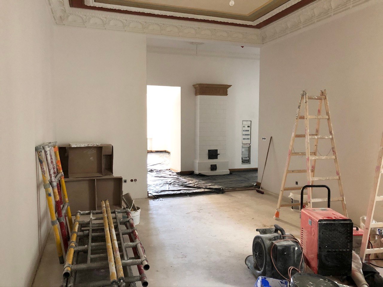 Wnętrze w trakcie remontu: widoczny piec kaflowy w sąsiednim pomieszczeniu, drabiny malarskie, osłonięta folią podłoga i narzędzia, ukazujące etap prac wykończeniowych.