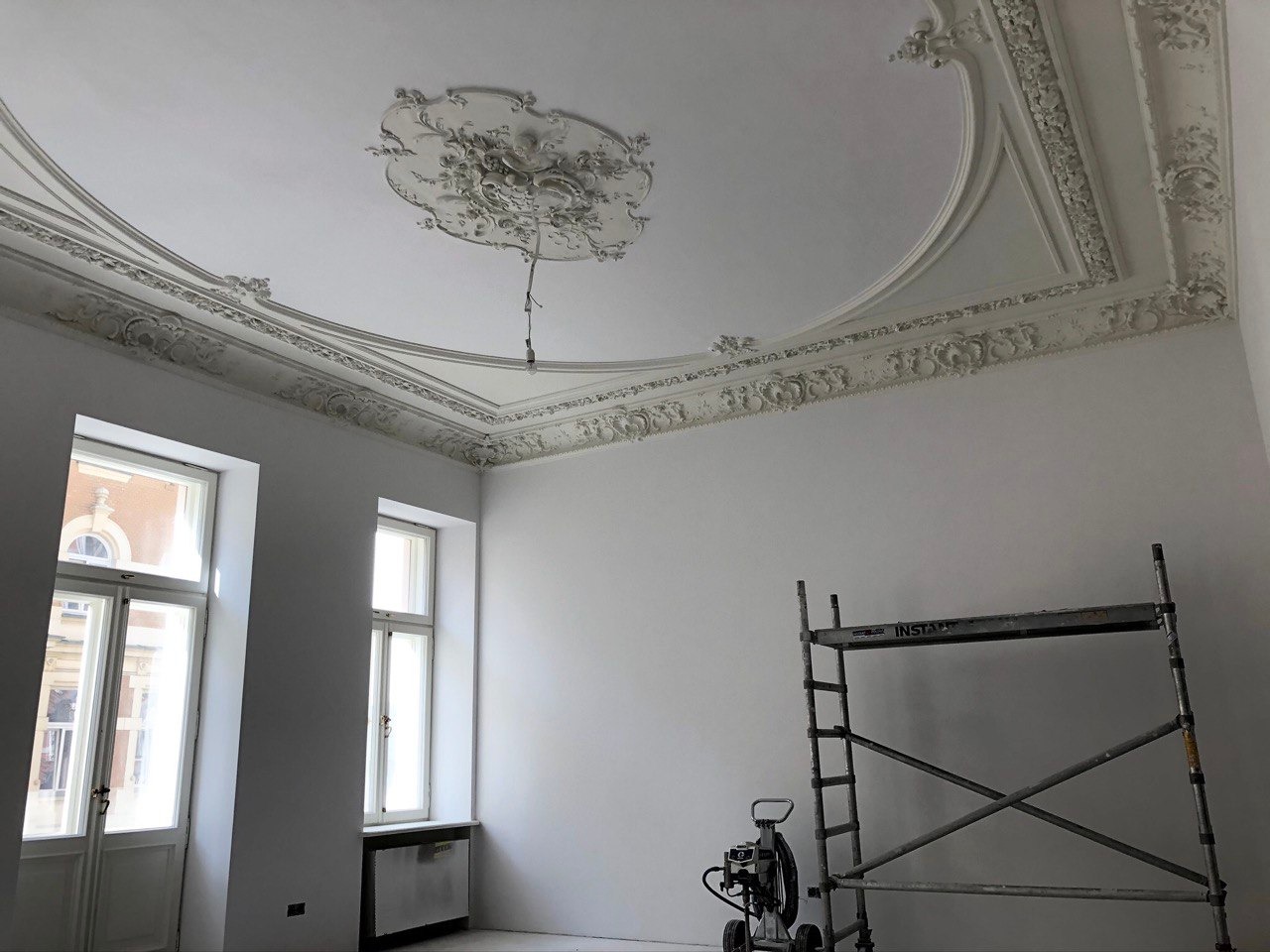 Pokój przed malowaniem: widoczne sztukaterie sufitowe, drabina i agregat malarski, dwa okna.