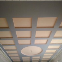 AndRom - Pomieszczenie z dekoracyjnym sufitem w odcieniach szarości i beżu, z widocznym oświetleniem i detalami sztukaterii.