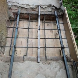 AndRom - Metalowa konstrukcja z prętów zbrojeniowych, prawdopodobnie pod balkon, przymocowana do zniszczonej betonowej płyty. Widoczne uszkodzenia betonu i cegieł.