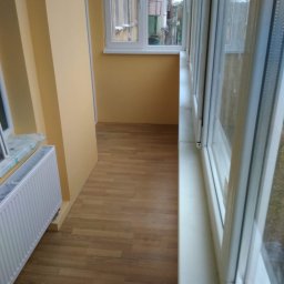 AndRom - Wykończony balkon z panelami podłogowymi, pomalowanymi na żółto ścianami i białą ramą okienną z widokiem na budynek mieszkalny.