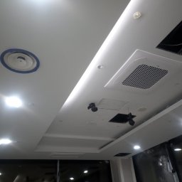 AndRom - Biały sufit podwieszany z zamontowaną klimatyzacją kasetonową LG, okrągłym anemostatem z niebieskim obramowaniem, oświetleniem punktowym LED i otworami rewizyjnymi w trakcie montażu.