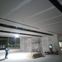 AndRom - Montaż płyt gipsowo-kartonowych na suficie w dużym pomieszczeniu biurowym, widoczne metalowe profile, pracownicy w kamizelkach odblaskowych i kaskach, rusztowanie oraz okna z widokiem na zewnątrz.