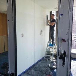 AndRom - Mężczyzna na aluminiowej drabinie w niebieskich ochraniaczach na buty szpachluje ścianę w jasnym pomieszczeniu biurowym z dużymi oknami, zabezpieczoną folią i taśmą malarską, widoczna szpachelka...