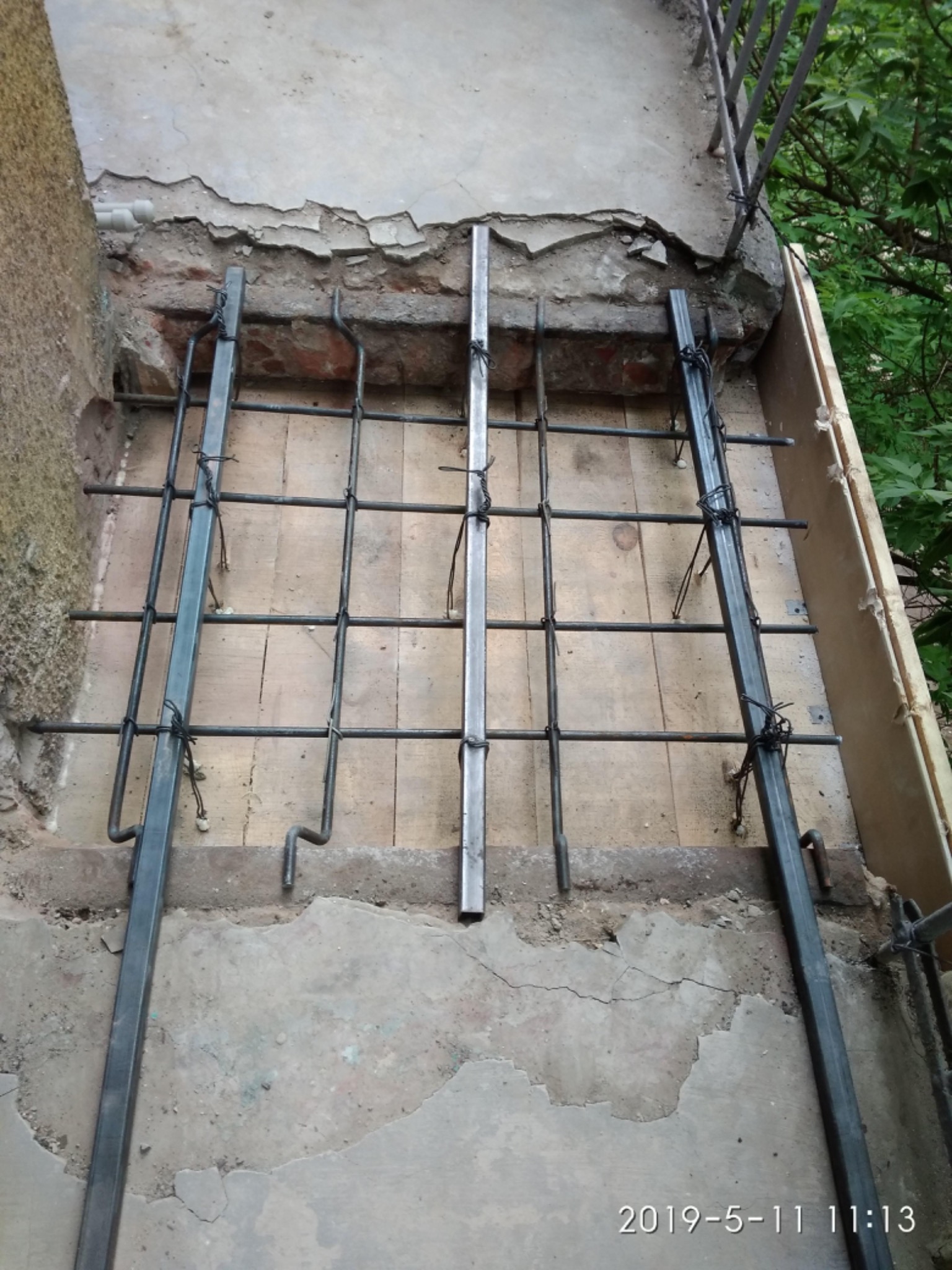 Metalowa konstrukcja z prętów zbrojeniowych, prawdopodobnie pod balkon, przymocowana do zniszczonej betonowej płyty. Widoczne uszkodzenia betonu i cegieł.