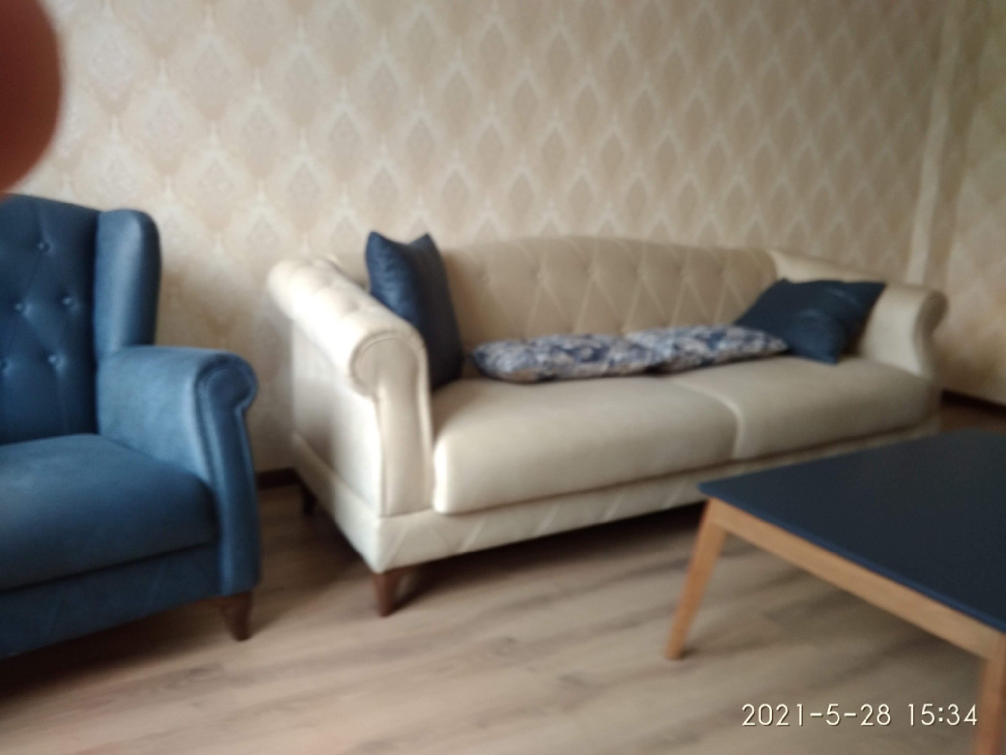 Umeblowane wnętrze w Warszawie: beżowa sofa z poduszkami, niebieski fotel i ciemny stolik kawowy na jasnej podłodze, ściany w delikatny wzór.