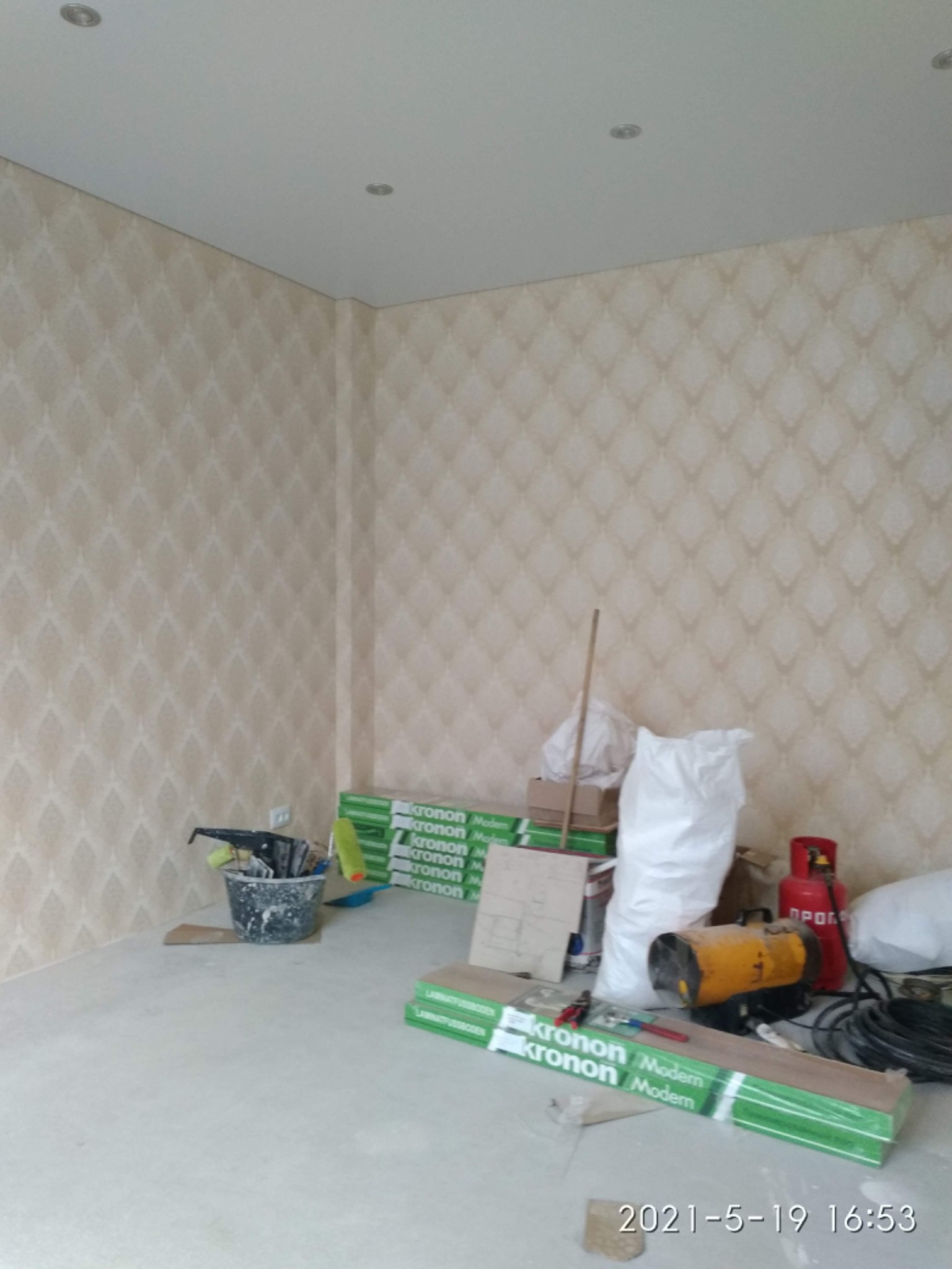 Pokój w trakcie prac wykończeniowych: ściany pokryte tapetą w delikatny wzór, widoczne narzędzia malarskie w wiadrze, paczki paneli podłogowych Kronon Modern, nagrzewnica, butla gazowa oraz worki...