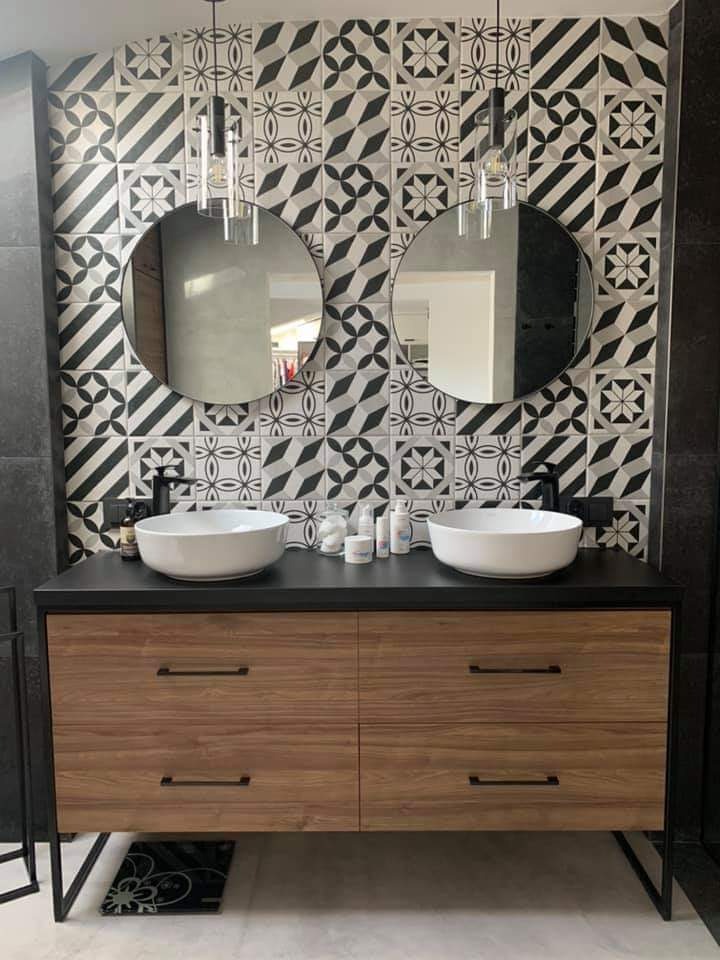 Nowoczesna łazienka z dwiema okrągłymi umywalkami nablatowymi, czarnym blatem i szafką z drewnopodobnym frontem, na tle ściany wyłożonej wzorzystymi, czarno-białymi płytkami.