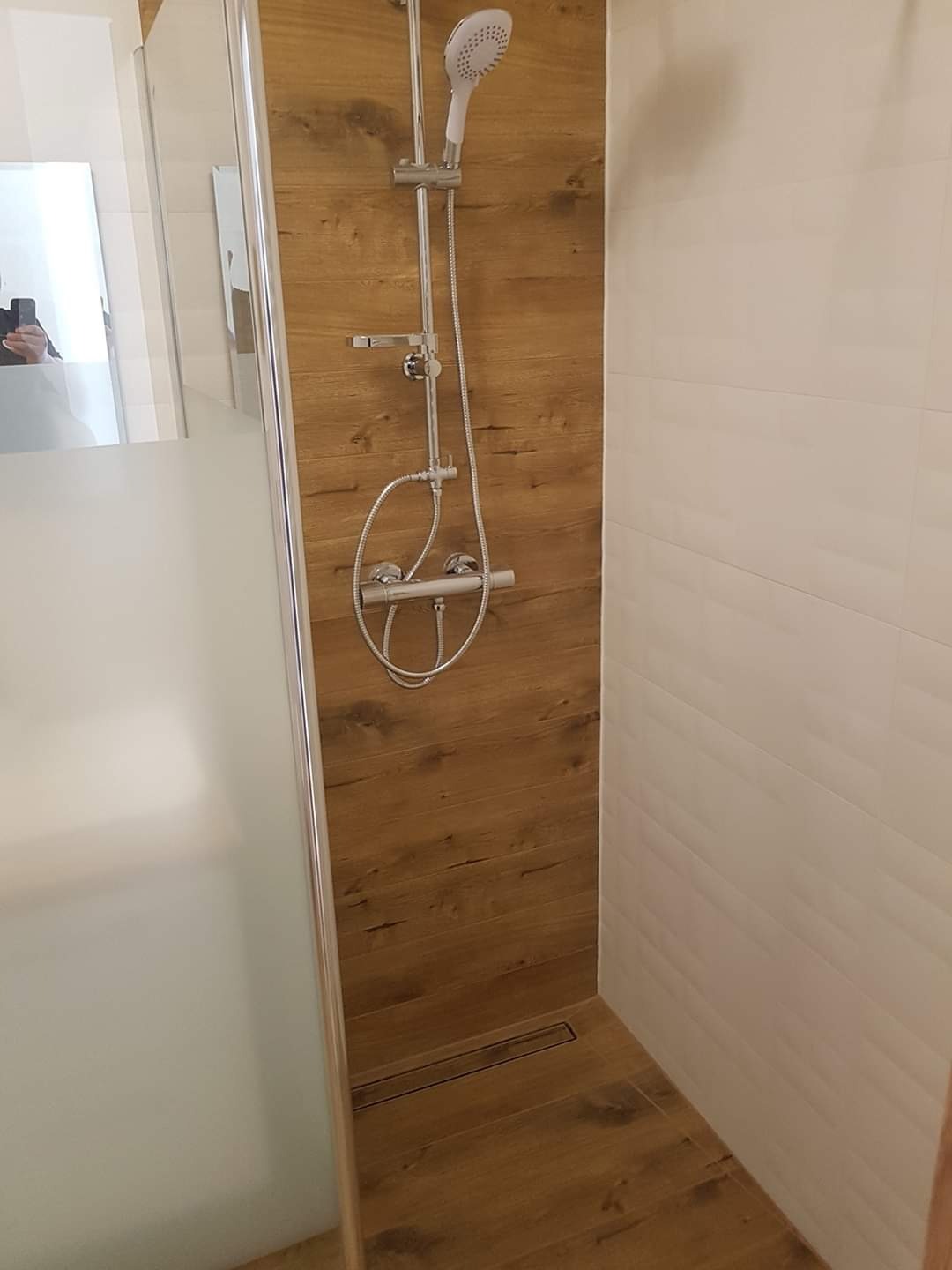 Nowoczesna kabina prysznicowa z deszczownicą, wykończona płytkami imitującymi drewno na ścianie i podłodze, oraz białymi, poziomymi płytkami na sąsiedniej ścianie. Widoczny odpływ liniowy w podłodze.