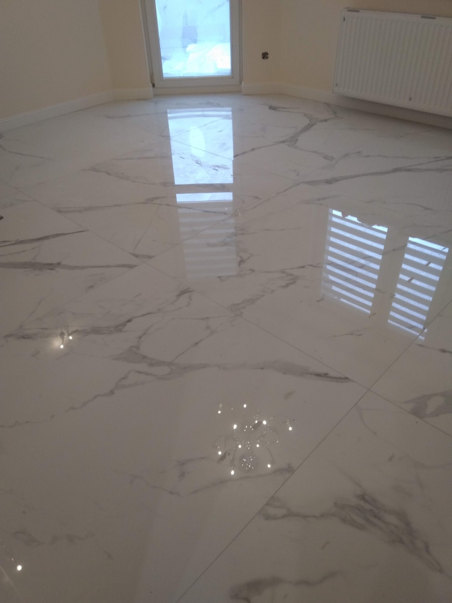 Błyszcząca, biała podłoga z płytek imitujących marmur, z widocznym odbiciem okna i rolet, oraz grzejnik na ścianie w tle.