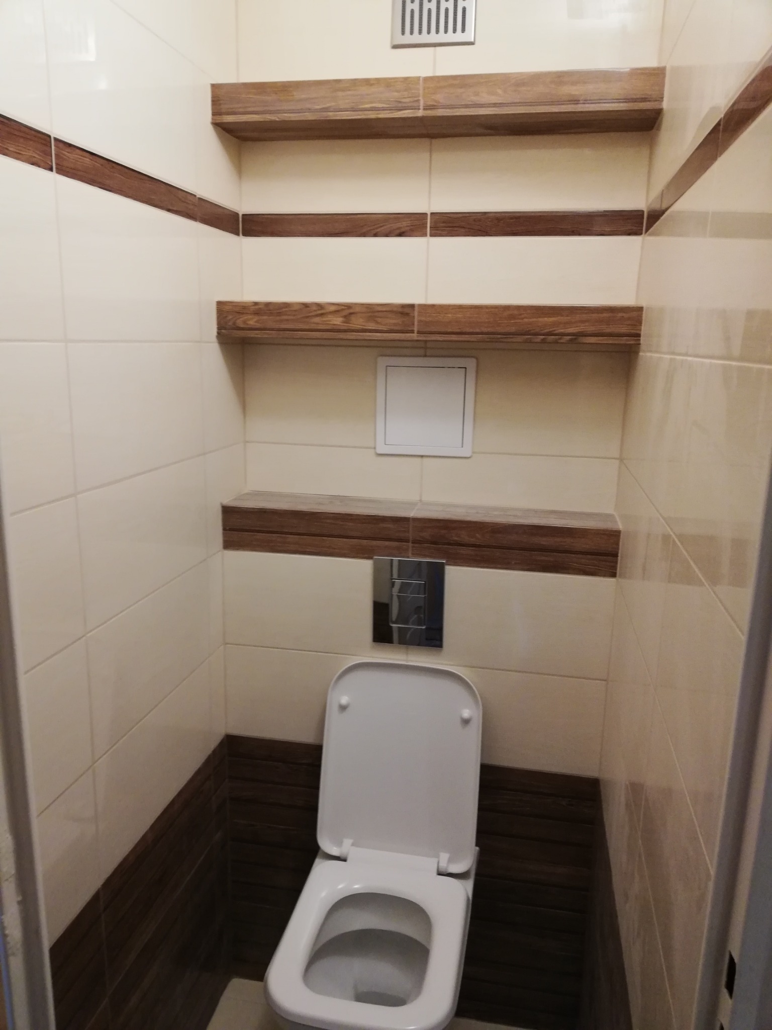 Wykończona łazienka z toaletą, jasnymi płytkami i drewnianymi akcentami w postaci półek i pasa dekoracyjnego.