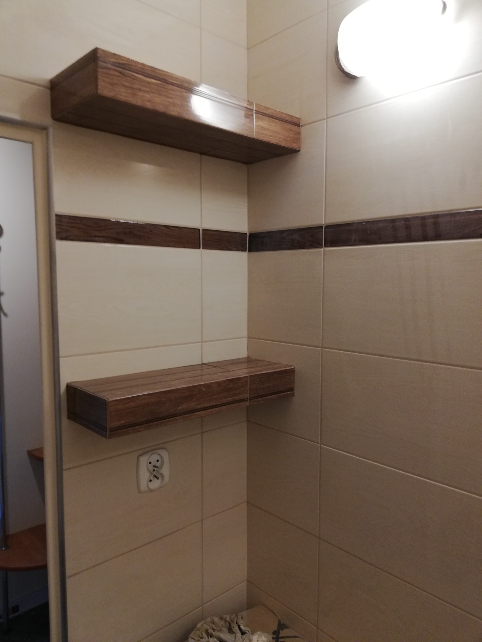 Dwie drewniane półki zamontowane na kafelkowanej ścianie w łazience z widocznym gniazdkiem elektrycznym i fragmentem drzwi.