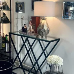 Konsola z czarną metalową podstawą o geometrycznym wzorze, szklanym blatem i dekoracjami w postaci lampy i kwiatów, ustawiona w eleganckim wnętrzu.