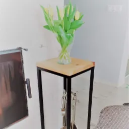 Stolik pomocniczy z drewnianym blatem i metalową ramą, na którym stoi szklany wazon z żółtymi tulipanami, obok znajduje się kominek z metalowymi akcesoriami