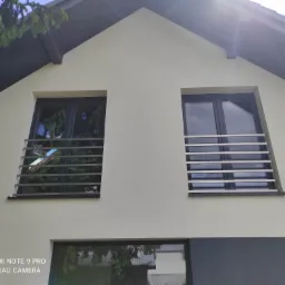 Elewacja domu z dwoma oknami zabezpieczonymi poziomymi, metalowymi prętami. Widoczne odbicia w szybach i fragment korony drzewa w górnym rogu.