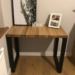 Biurko z blatem z klejonego drewna i czarnymi, metalowymi nogami, dekorowane ramką ze zdjęciami i obrazem z motywem roślinnym.