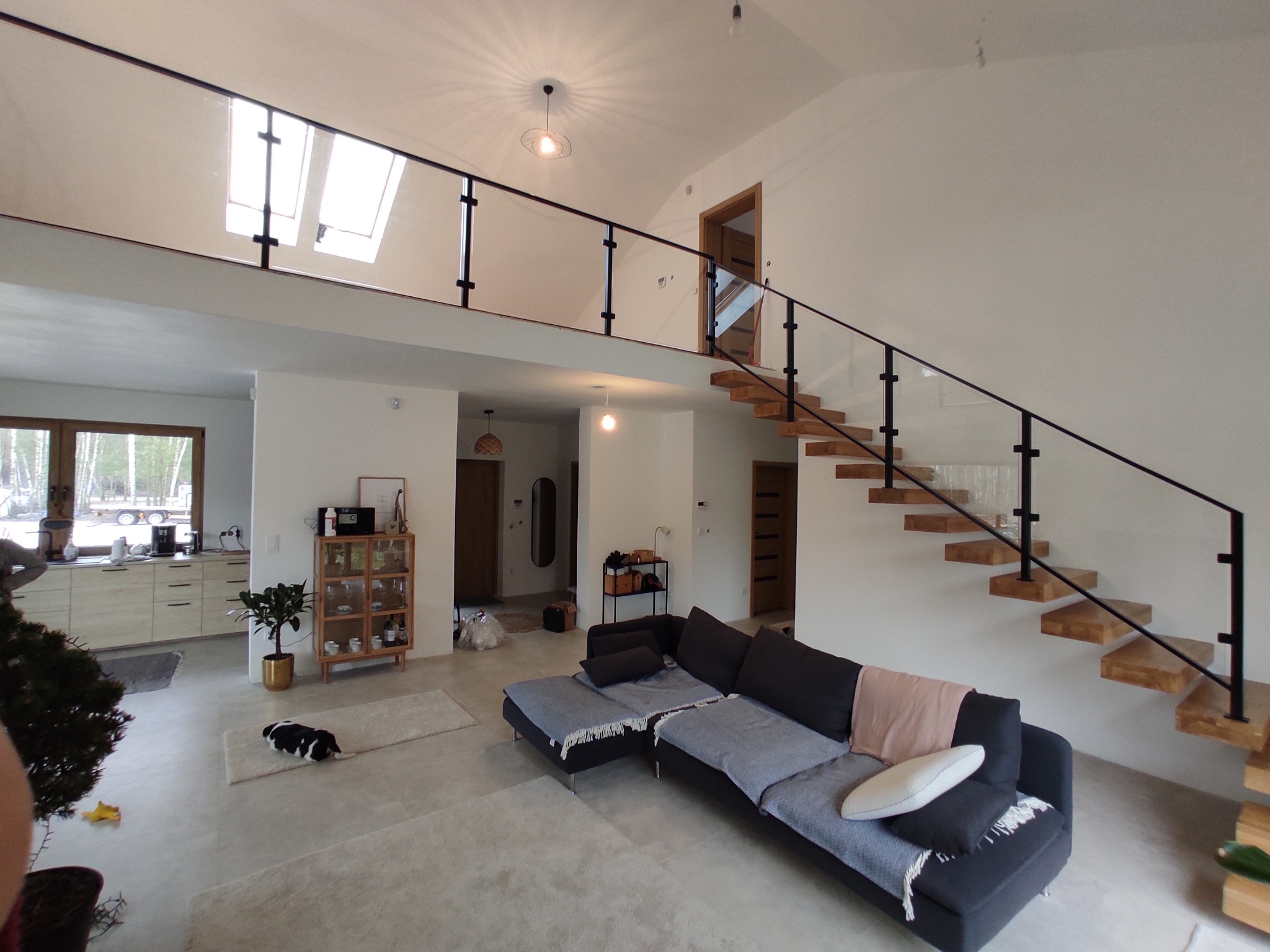 Nowoczesne wnętrze domu z drewnianymi schodami wspornikowymi z czarną balustradą, szklanymi panelami i czarnymi słupkami. Na górze balustrada z czarnymi słupkami i szklanymi panelami. W tle...