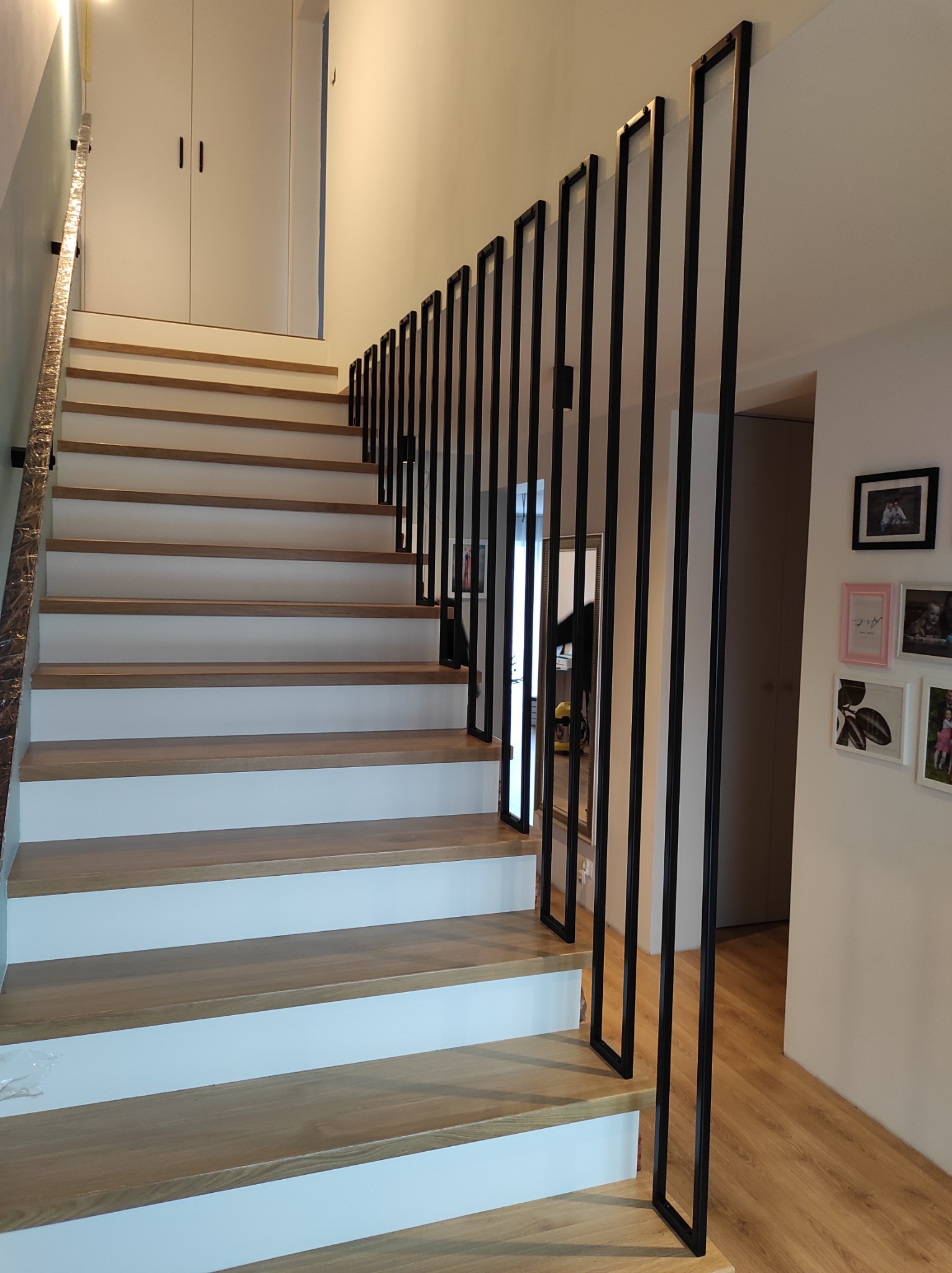 Nowoczesne schody drewniane z minimalistyczną balustradą z pionowych, czarnych, metalowych profili o kwadratowym przekroju, na tle białych ścian i drewnianej podłogi.
