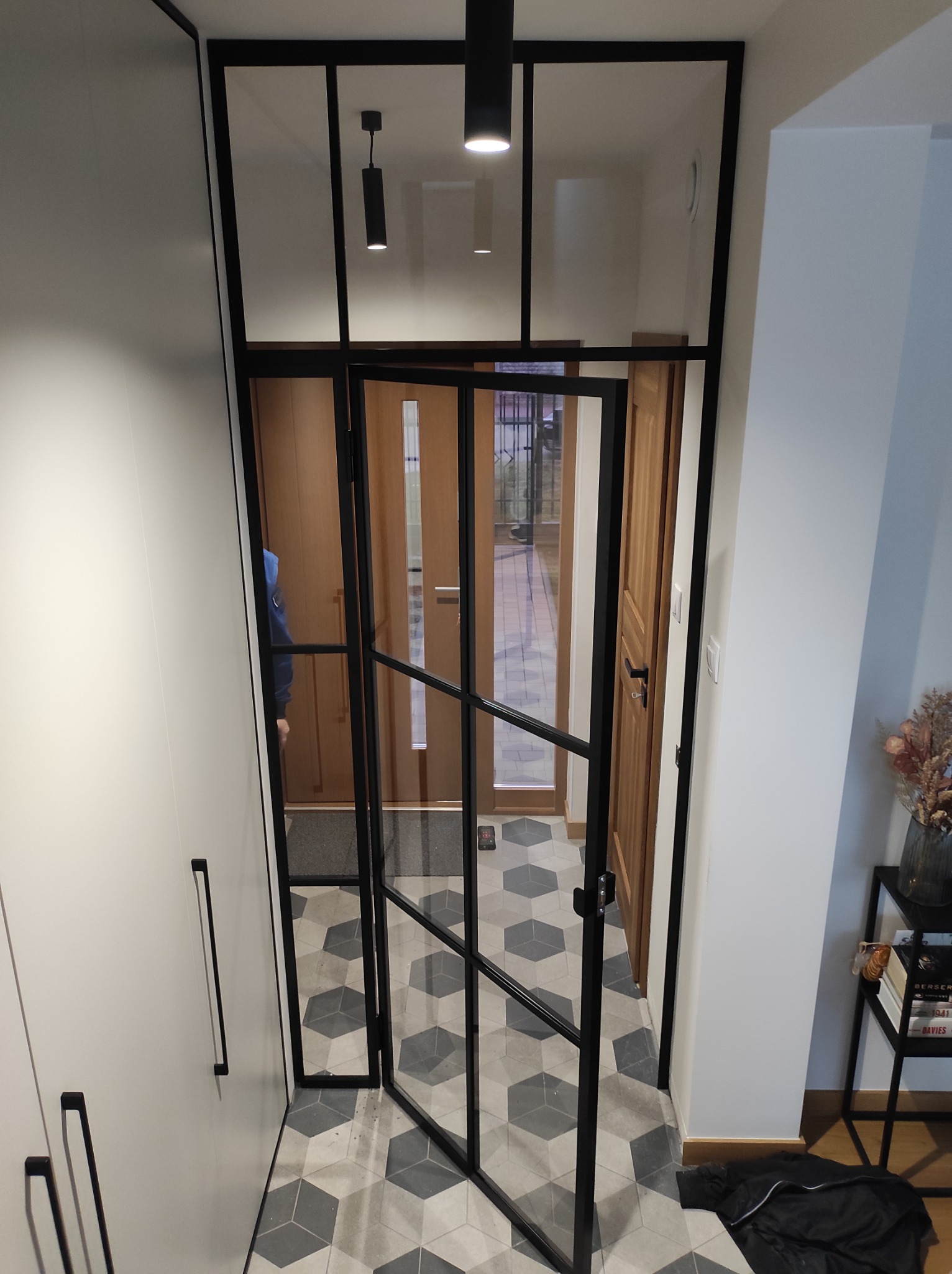 Wnętrze przedpokoju z drzwiami w metalowej ramie z szybami, otwarte częściowo, widoczna podłoga w geometryczny wzór i elementy wyposażenia.