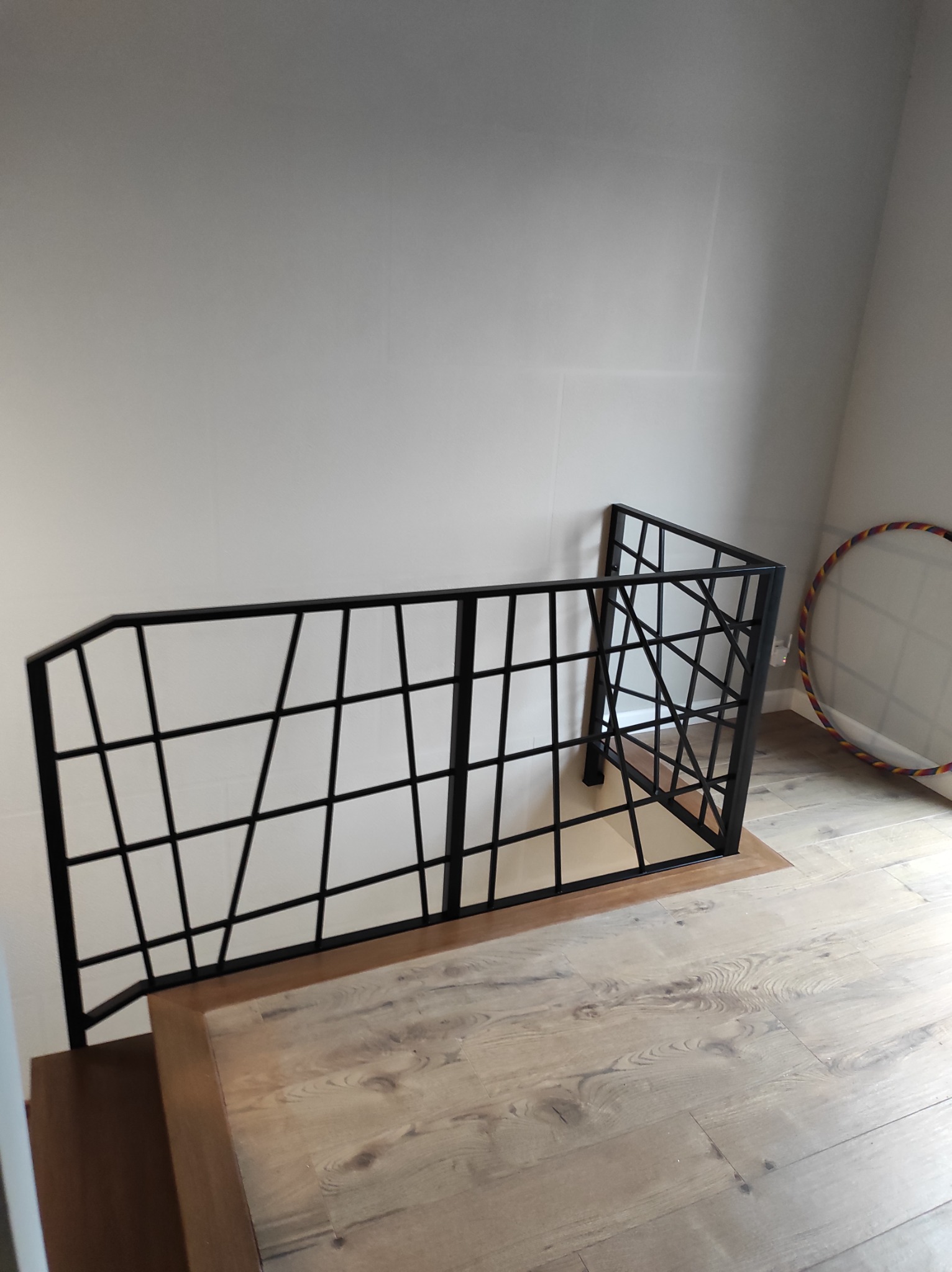 Nowoczesna, czarna barierka schodowa z metalowych profili o geometrycznym wzorze, zamontowana przy zejściu na jasnych drewnianych schodach, widoczny fragment podłogi i ściany w tle.