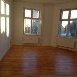 Pusty pokój po remoncie z odnowioną drewnianą podłogą, świeżo pomalowanymi ścianami i dwoma oknami wpuszczającymi jasne światło słoneczne.