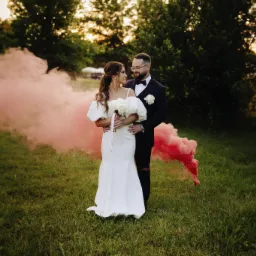 Para młoda w objęciach na łące, otoczona różową mgłą z dymnych świec. Panna młoda w białej sukni, pan młody w garniturze. Romantyczna sesja plenerowa.